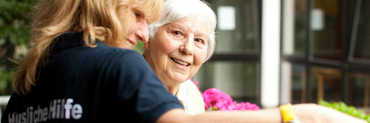 Eine lachende Seniorin mit einer DRK-Mitarbeiterin beim Blumen gießen. 